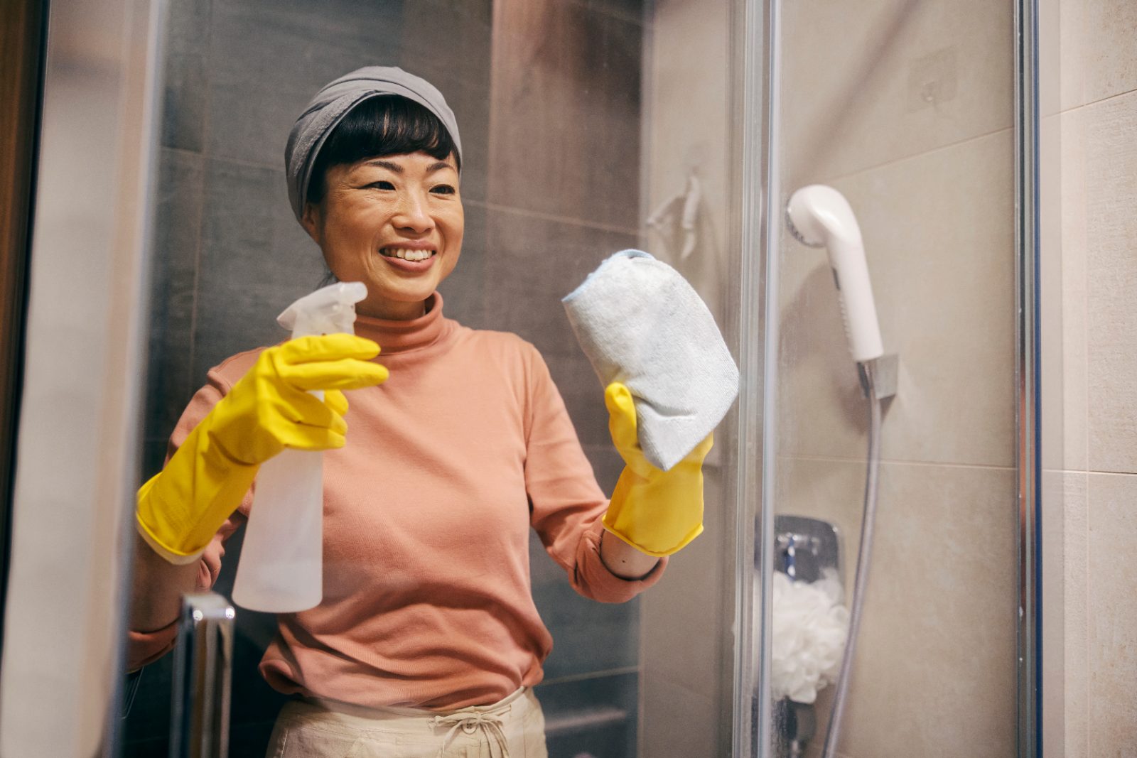 A woman in rubber gloves cleaning the glass screen of a bathroom shower