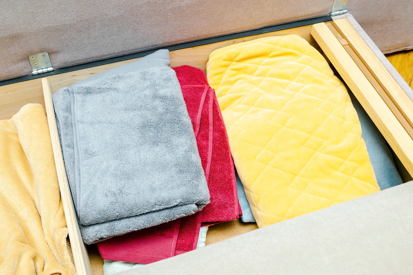 An open storage drawer under a wooden bed frame, packed tightly with folded towels and blankets