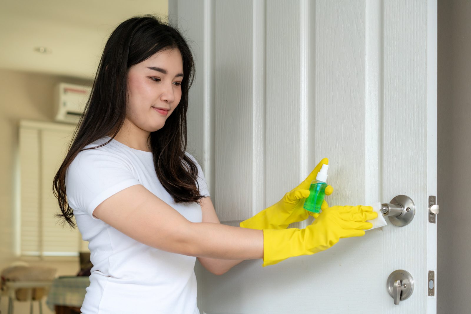 A woman in yellow gloves wiping a door handle with a cloth and spray bottle in a bright home