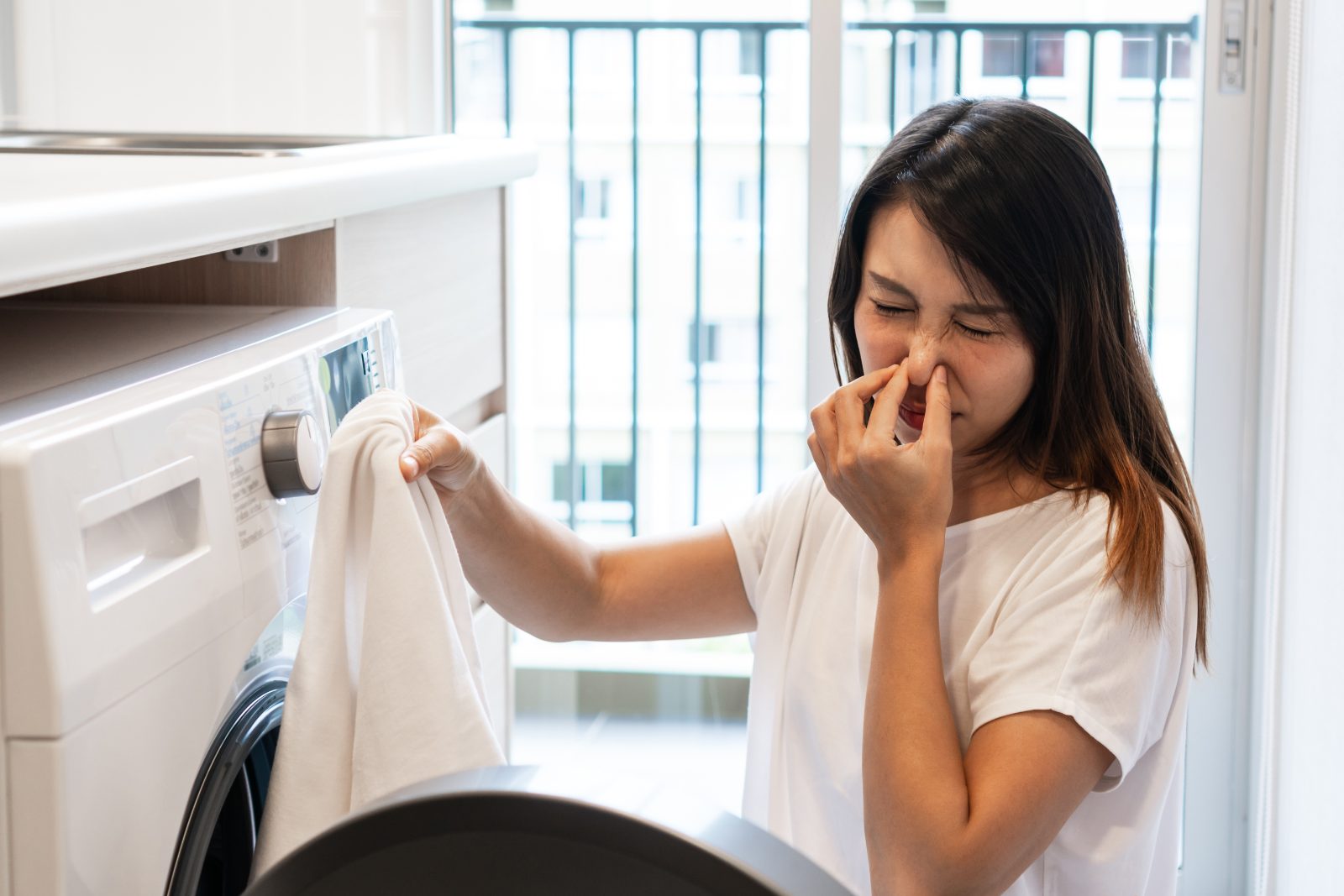 Woman pulling laundry from a front-loading washing machine in a service yard and reacting to the smell