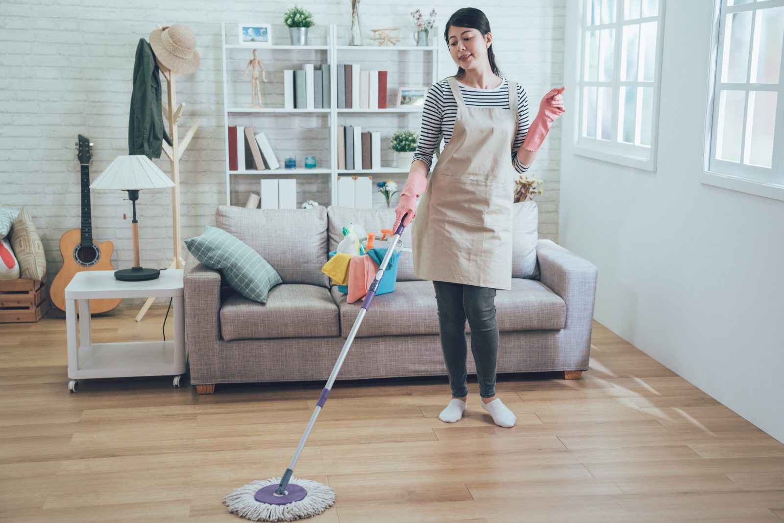 A young woman in an apron and pink rubber gloves mopping the floor of a bright apartment living room