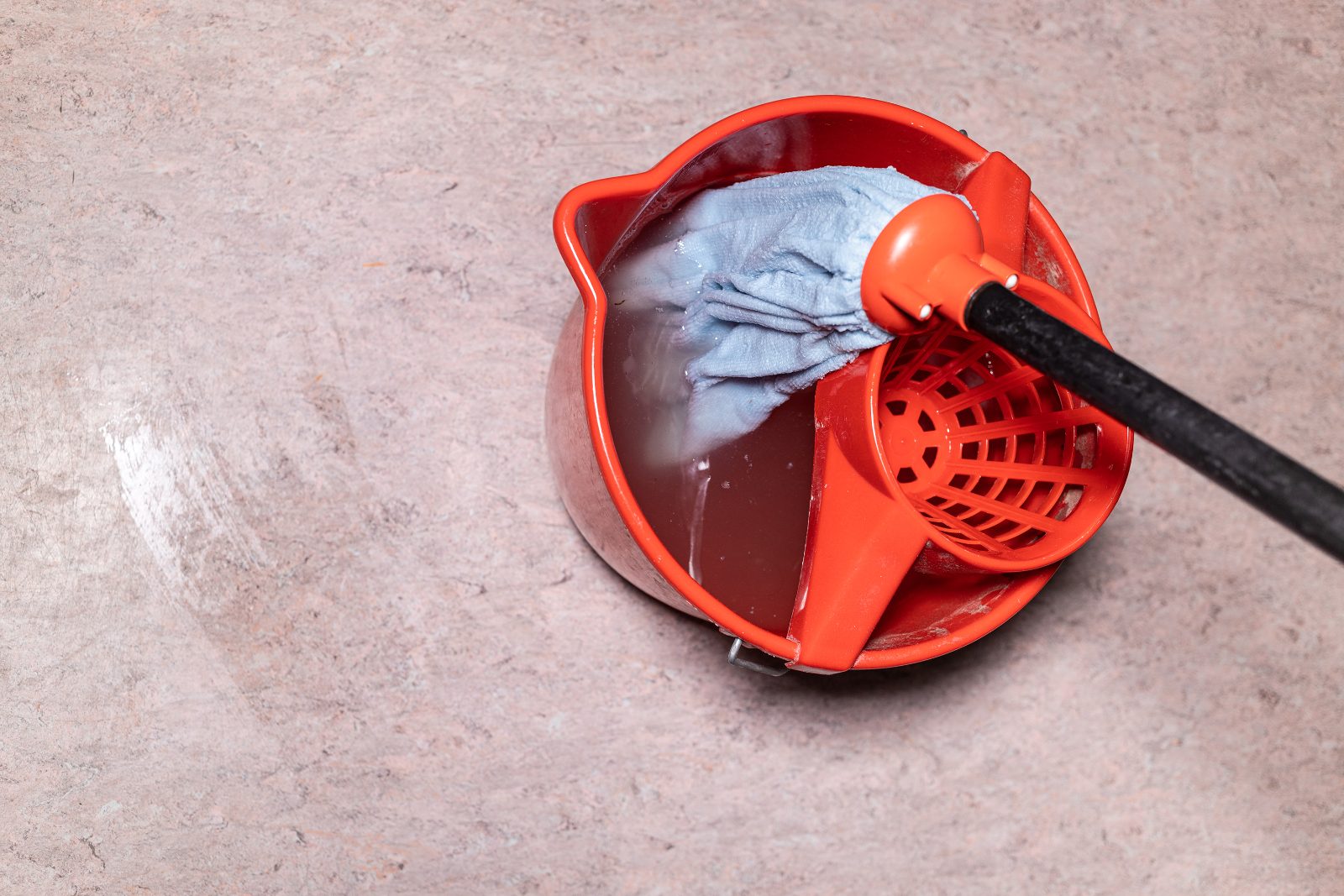 Top view of a mop resting in a red bucket of brown, dirty water on a tiled floor