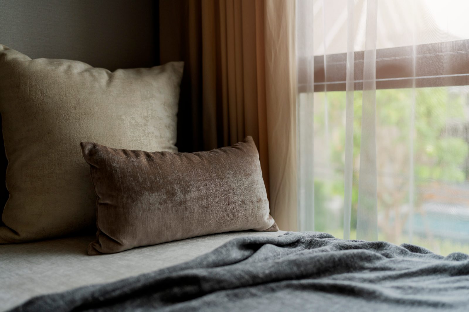 Velvet cushions on a bed beside a sheer curtain and a window screen with greenery outside