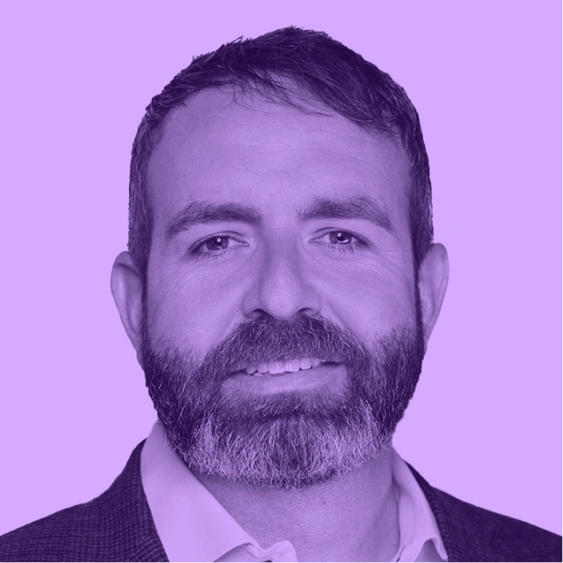 Portrait of a person with short hair and a beard, wearing a collared shirt and jacket, on a purple background