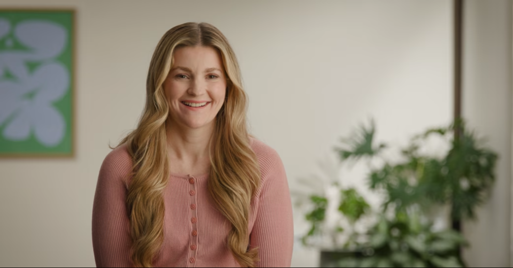 Smiling woman with long wavy blonde hair wearing a pink buttoned sweater, sitting indoors with plants and abstract art in the background.