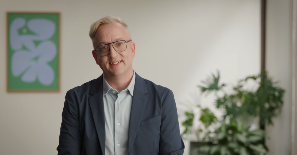 Smiling man with blonde hair and glasses wearing a navy blazer and light blue shirt standing indoors with blurred green plants and framed abstract art in the background.