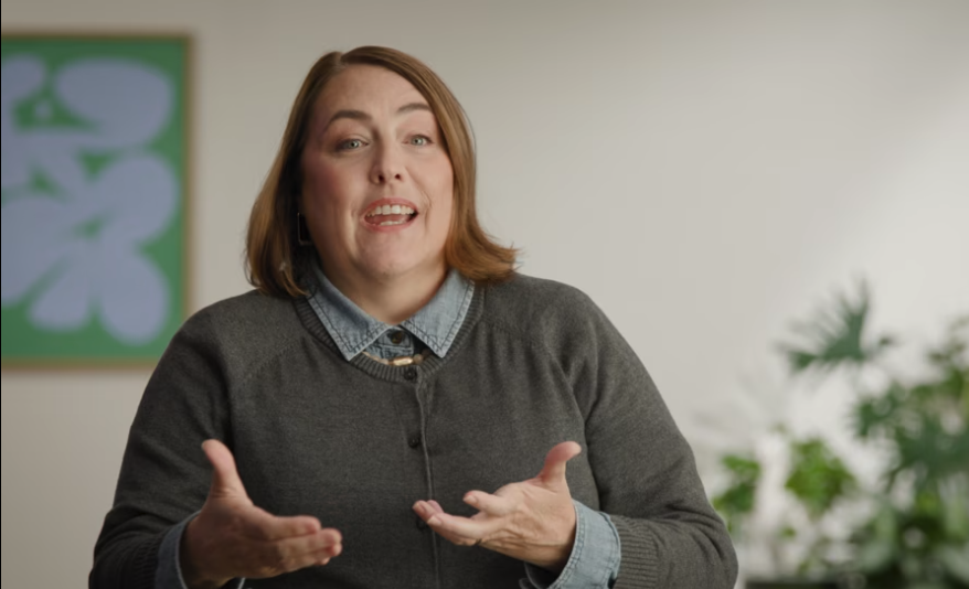 Woman with shoulder-length brown hair wearing a gray sweater over a denim shirt gesturing with her hands while speaking.