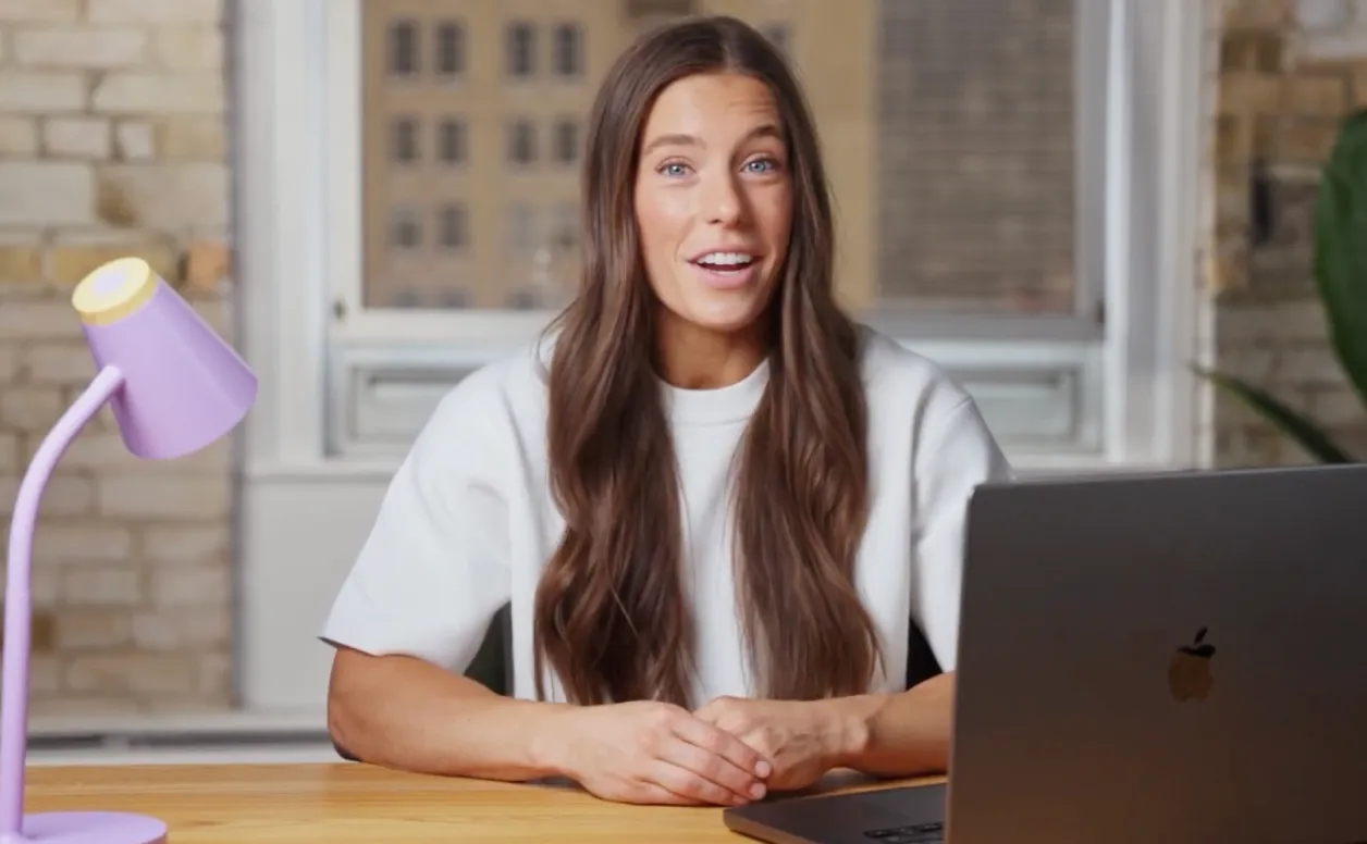 A woman with long brown hair wearing a white shirt sits at a wooden desk with a laptop open in front of her and a purple desk lamp on the left side, speaking and smiling, with a brick wall and window in the background.