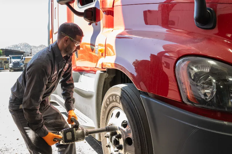 Mechanic using impact wrench to remove lug nuts from semi-truck wheel, wearing gloves and safety glasses.