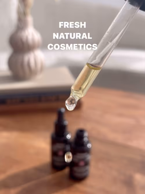 Eine Pipette mit einem Tropfen Öl, im Hintergrund zwei unscharfe Flaschen auf einem Holztisch und der Text 'FRESH NATURAL COSMETICS'.