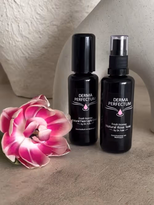 Zwei schwarze Flaschen mit der Aufschrift Dermaperfectum©, ein natürlicher Gesichtscreme- und Rosen-Toniktropfen, neben einer pink-weißen Blume auf einem grauen Untergrund.
