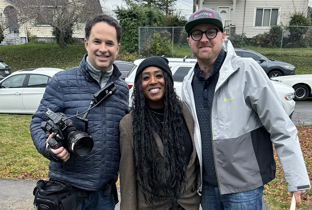 Three people standing outside smiling; the man on the left is holding a professional video camera.