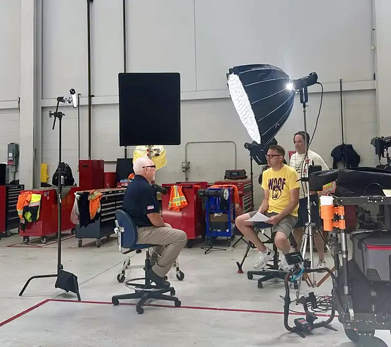 Two men sitting on rolling chairs facing each other in a workshop, surrounded by professional video equipment and toolboxes.