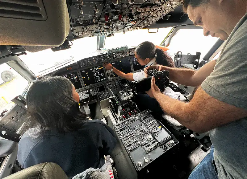 Two women, an instructor and a student, sitting in an airplane cockpit while a man films their conversation.
