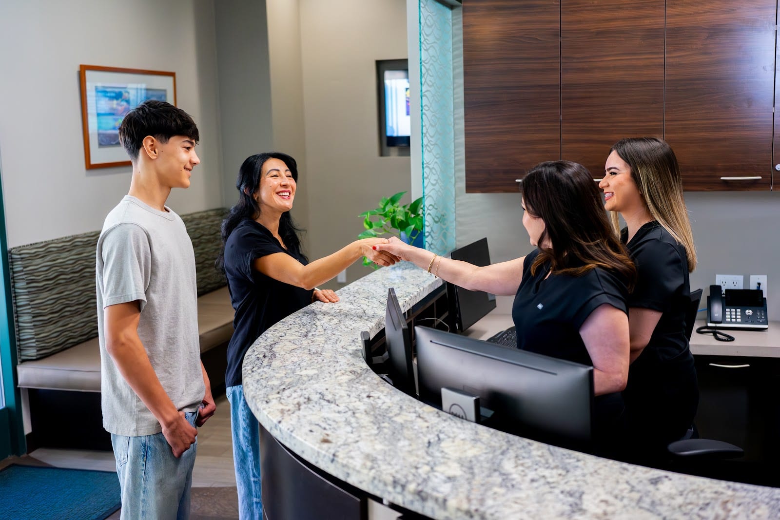 Two dental office staff members in black uniforms greet two patients with a handshake at the granite reception desk.