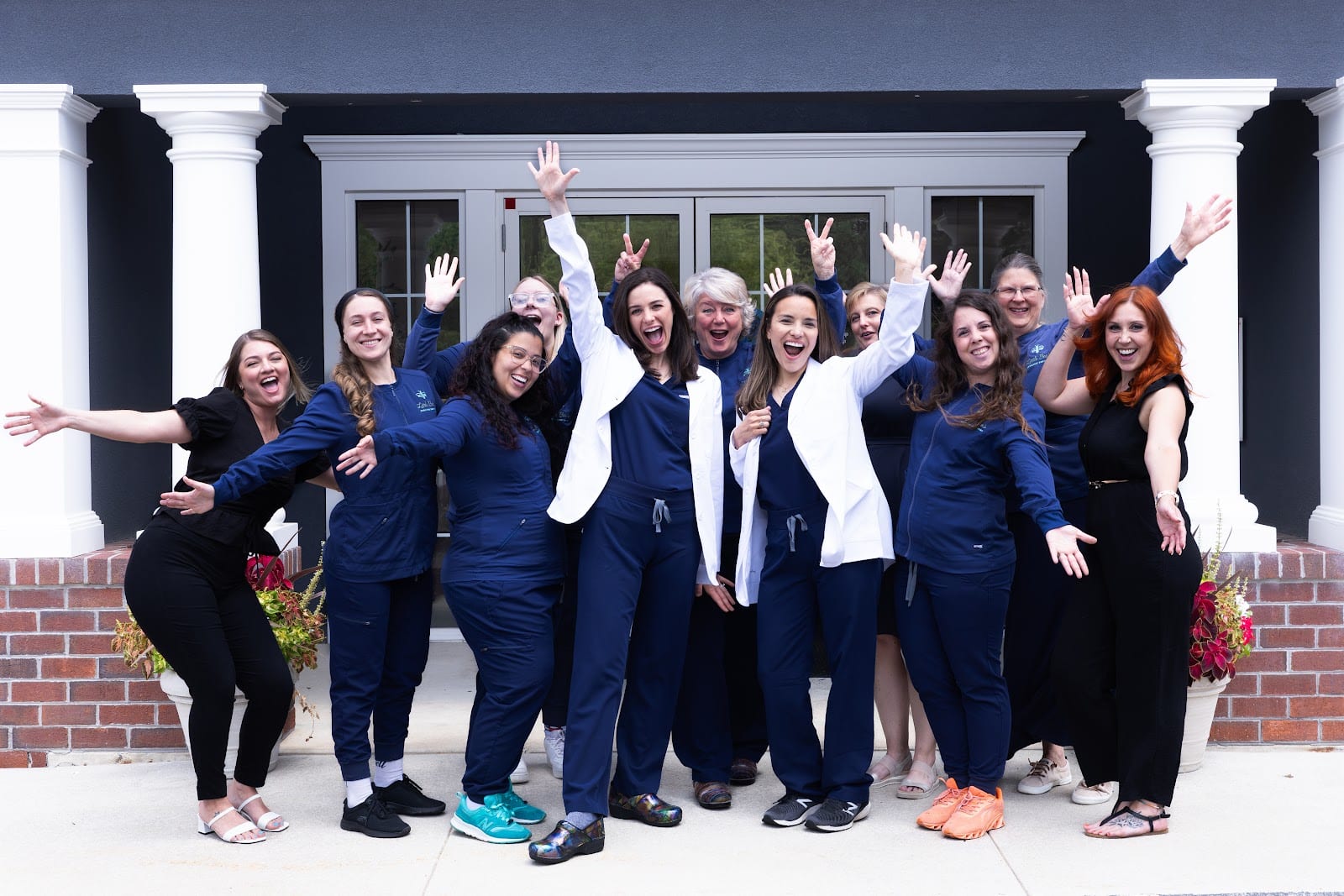 Twelve excited female dental staff members laughing and posing outside the dark blue practice exterior.