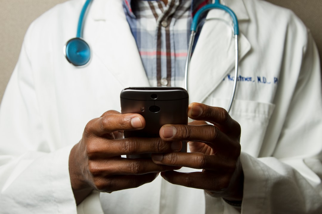 A doctor in a white coat with a stethoscope using a smartphone to message a patient — Photo by National Cancer Institute on Unsplash