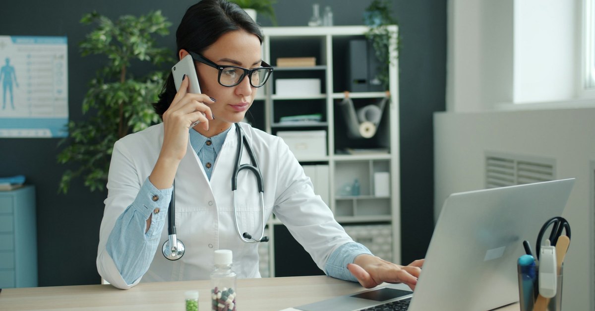 Clinic doctor confirming appointments on a smartphone next to her laptop