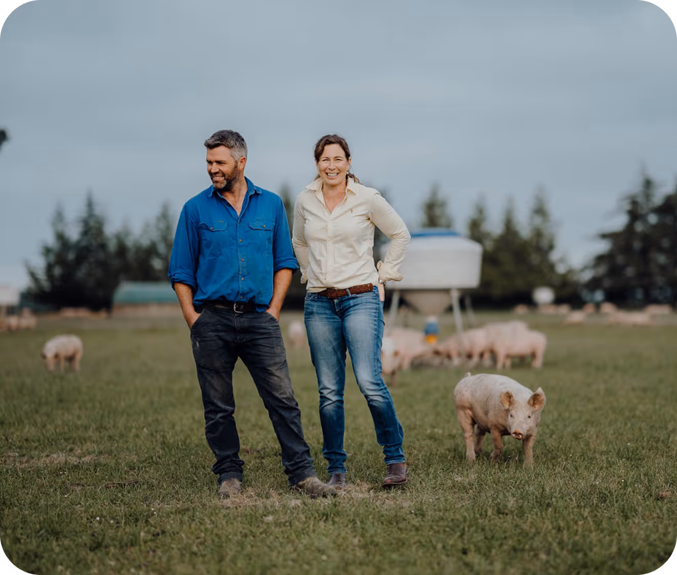 Smiling farmers standing in a field with free-range pigs grazing in the background.
