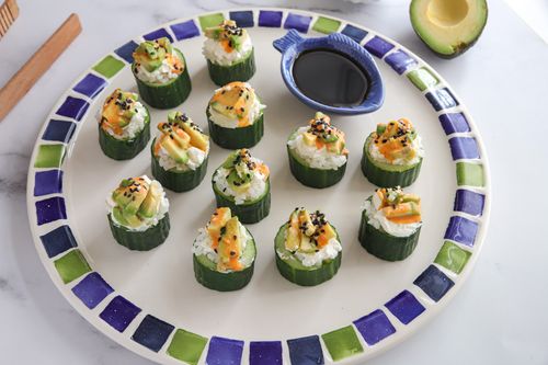Close-up of cucumber & avocado sushi bites topped with cream cheese, sesame seeds, and mayo, served on a wooden board with soy sauce — an easy no-roll sushi snack.