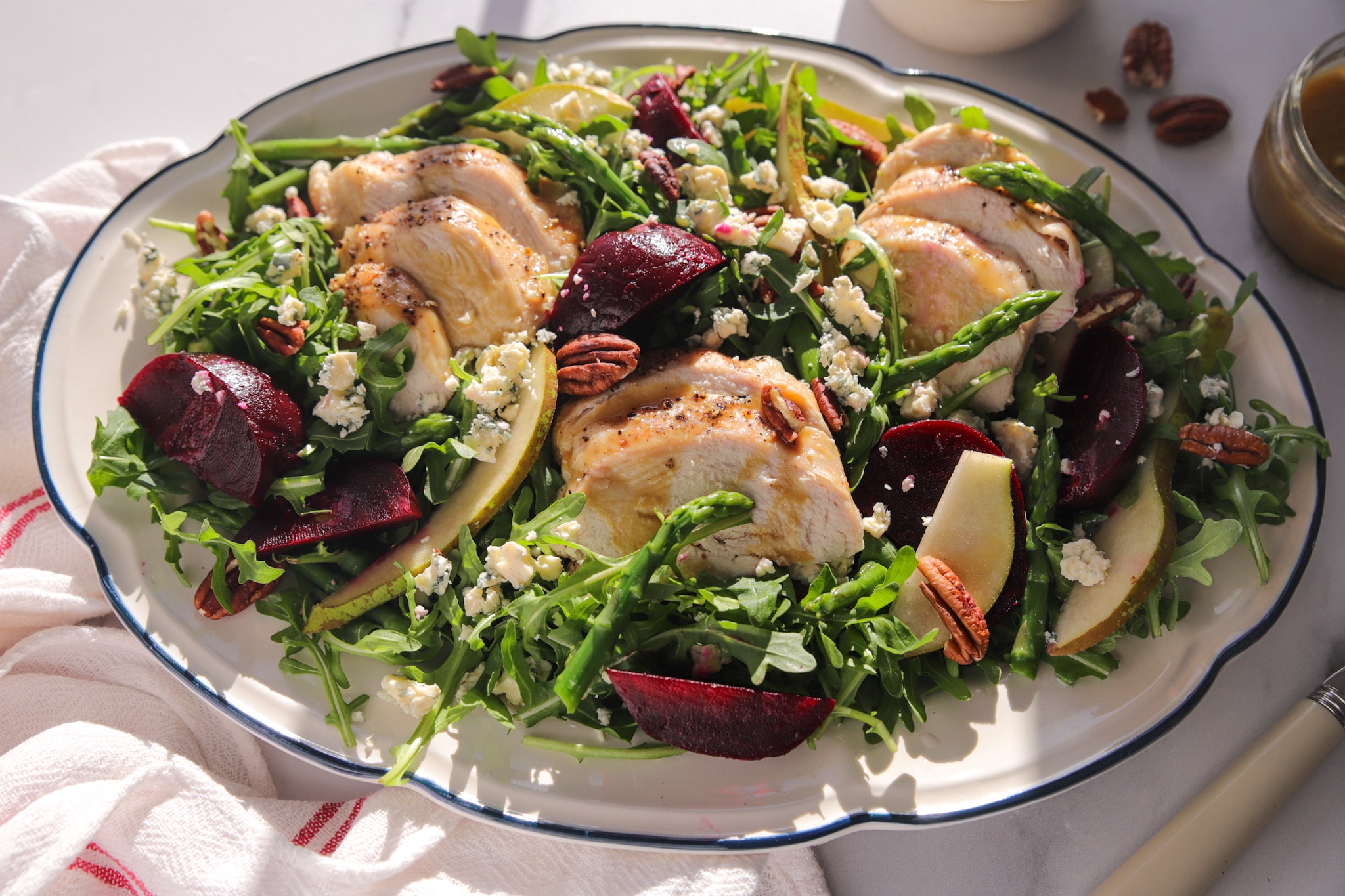 A colourful seasonal salad with sliced chicken breast, roast beetroot wedges, asparagus, pear, pecans and blue cheese on a white plate.