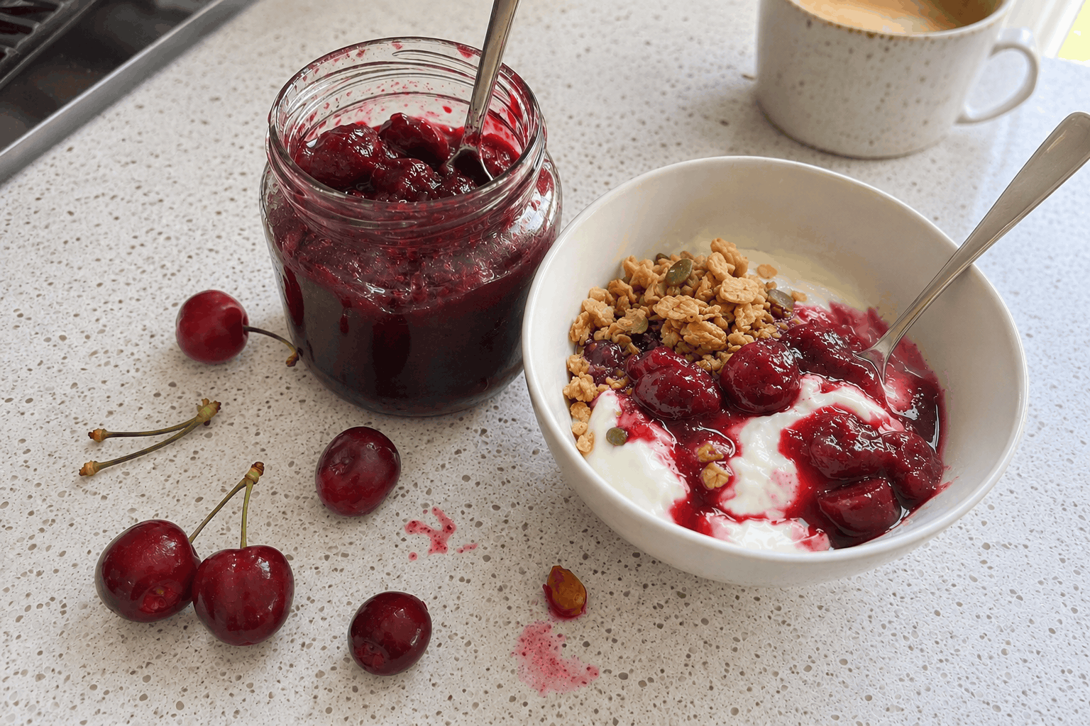 Bowl of Greek yoghurt with granola and homemade cherry compote, next to a jar of compote and fresh cherries on a white kitchen benchtop