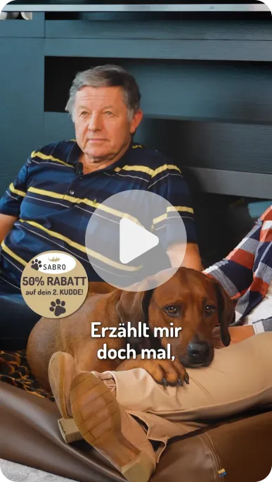 Älterer Mann in gestreiftem Hemd sitzt auf Sofa neben braunem Hund, der den Kopf auf einem Bein liegen hat.