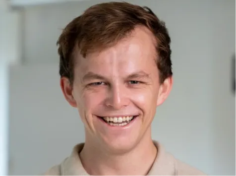 Lächelnder junger Mann mit braunen Haaren in einem beigen Hemd vor unscharfem Hintergrund.