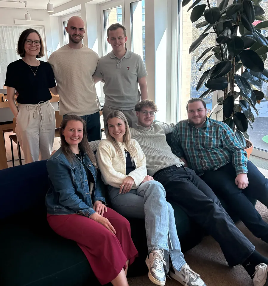 Sieben Personen, drei stehend und vier sitzend auf einem Sofa in einem hellen Büro, lächeln in die Kamera.