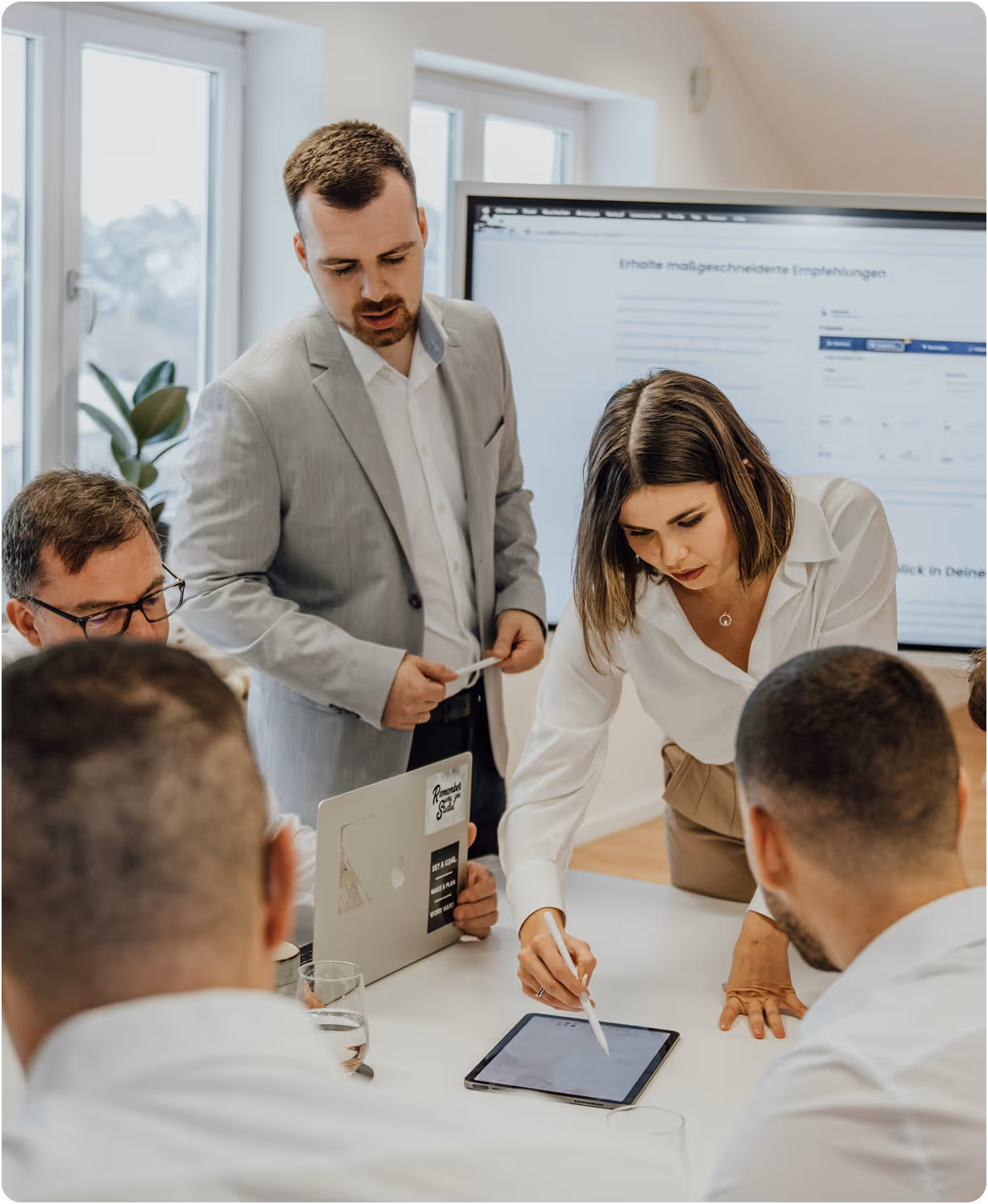 Team arbeitet in einem modernen Büro zusammen, eine Frau zeigt etwas auf einem Tablet, während Kollegen aufmerksam zuhören.
