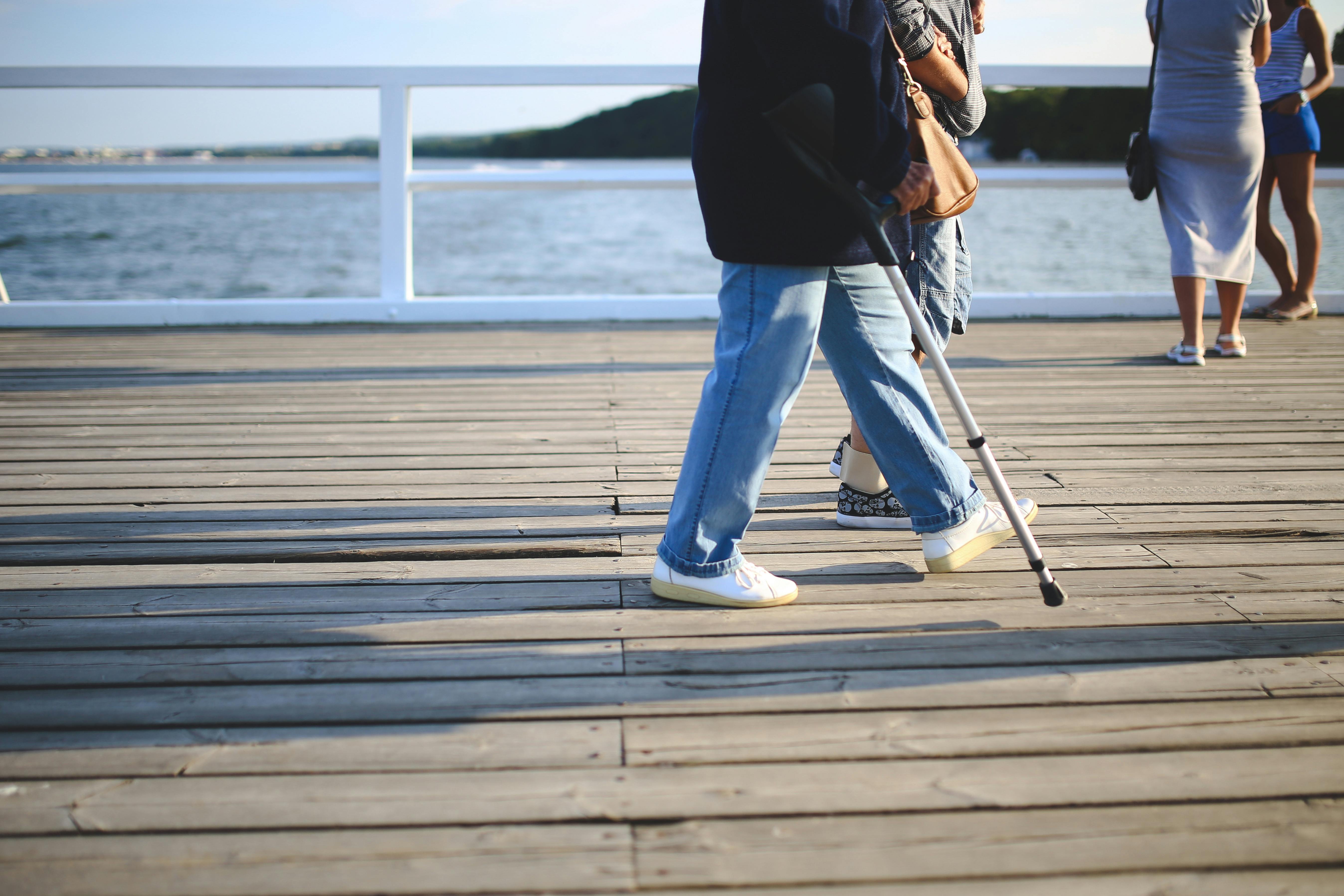 Injured person walking with a crutch on a waterfront boardwalk