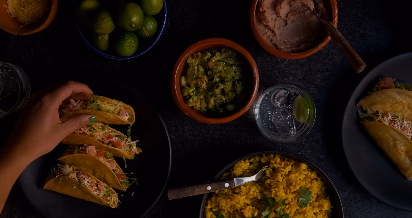 Low Transparency Hand reaching for freshly made crunchy taco at catered Mexican dinner table with yellow rice, beans, guacamole, and lime