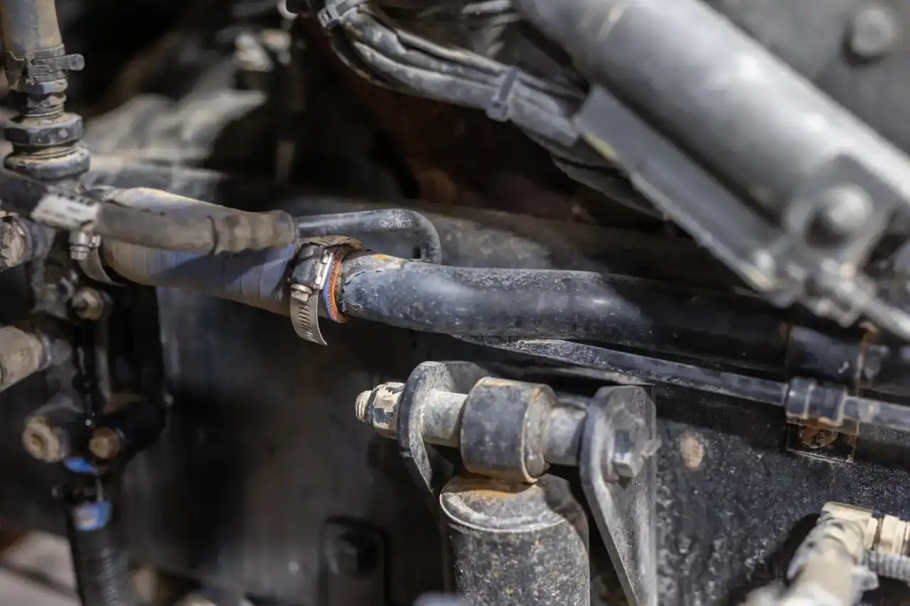 Hydraulic hose secured with steel worm-drive clamp amid dusty loader linkage, illustrating maintenance inspection focus.