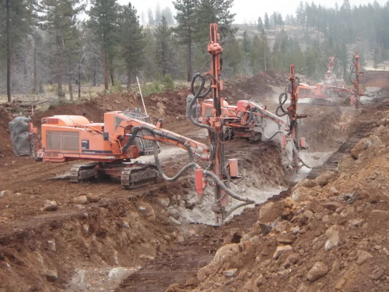 Heavy-duty drilling service uses multiple track-mounted rock drills trenching soil at a construction site in wooded terrain.