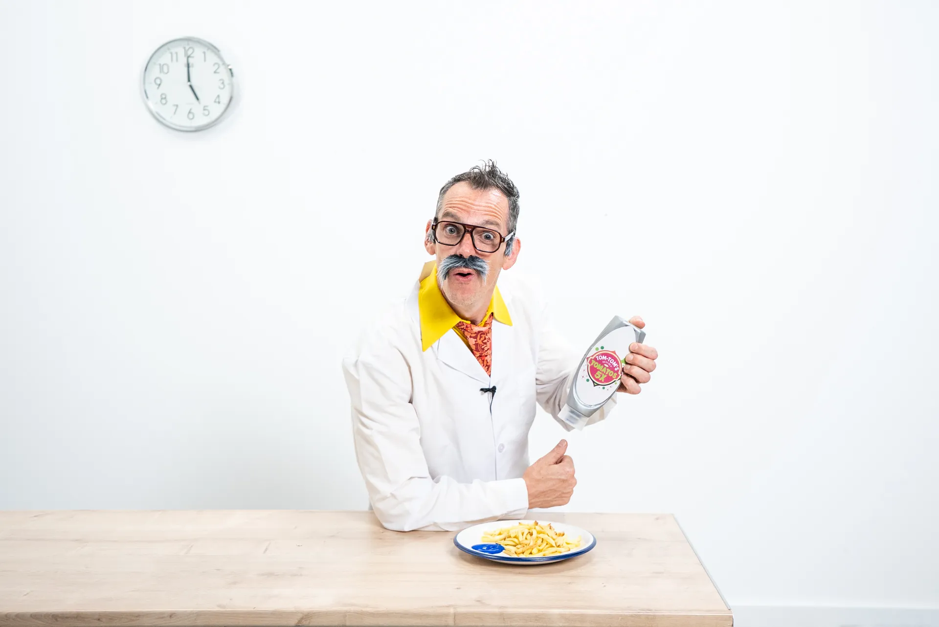 Man with a fake mustache and glasses sitting at a table holding a ketchup bottle labeled 'Tom-Tom Tomato 51%' next to a plate of fries with blue sauce.