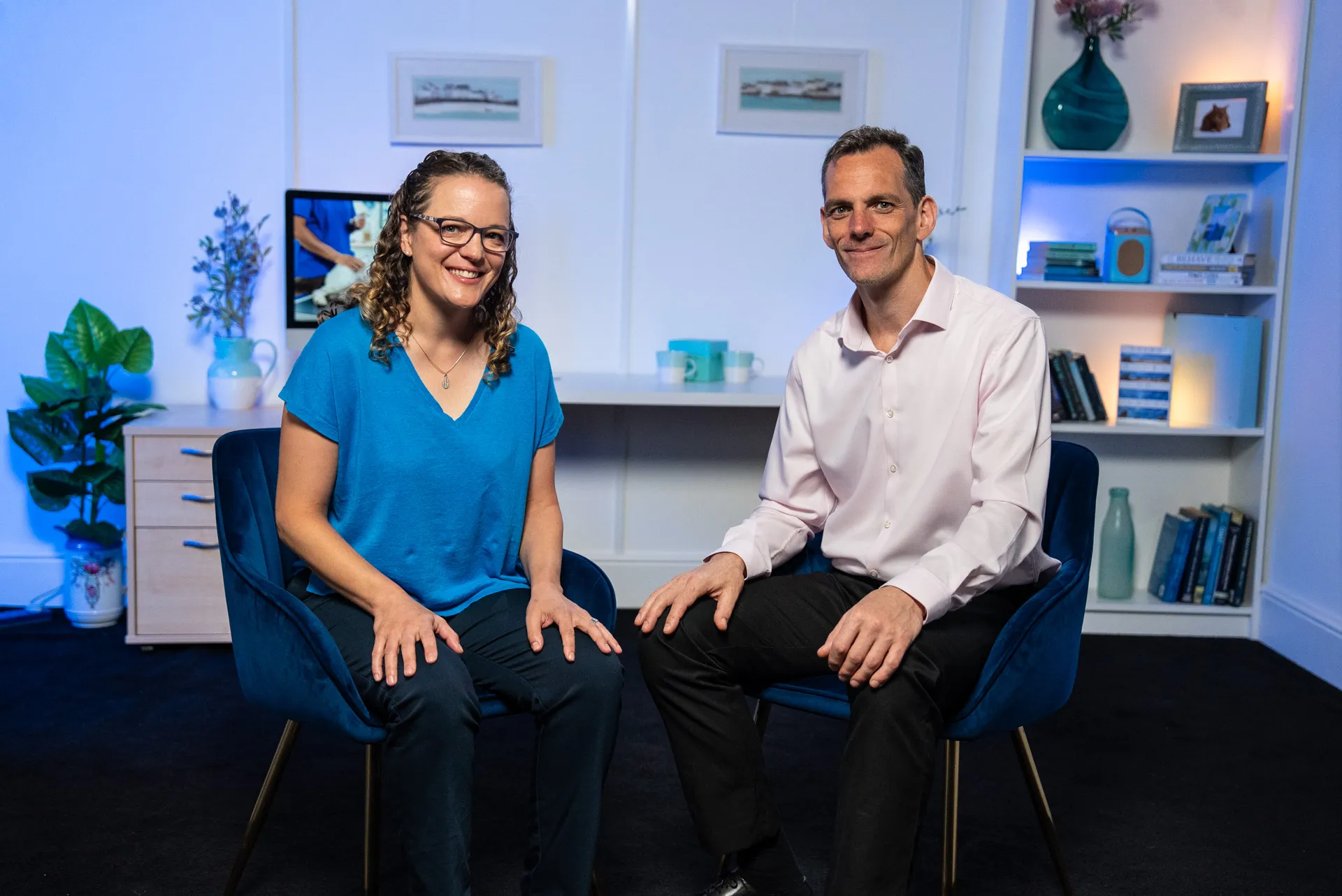 Woman in a teal blouse animatedly talking to a man in a navy scrub top in an office setting with pet posters in the background.
