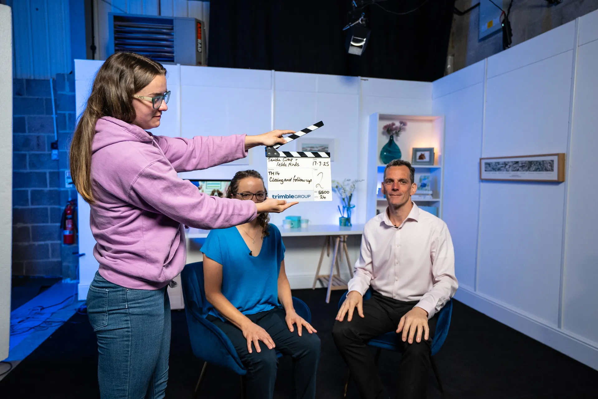 Woman in purple hoodie holding a film clapperboard in front of two seated people in a studio setting.