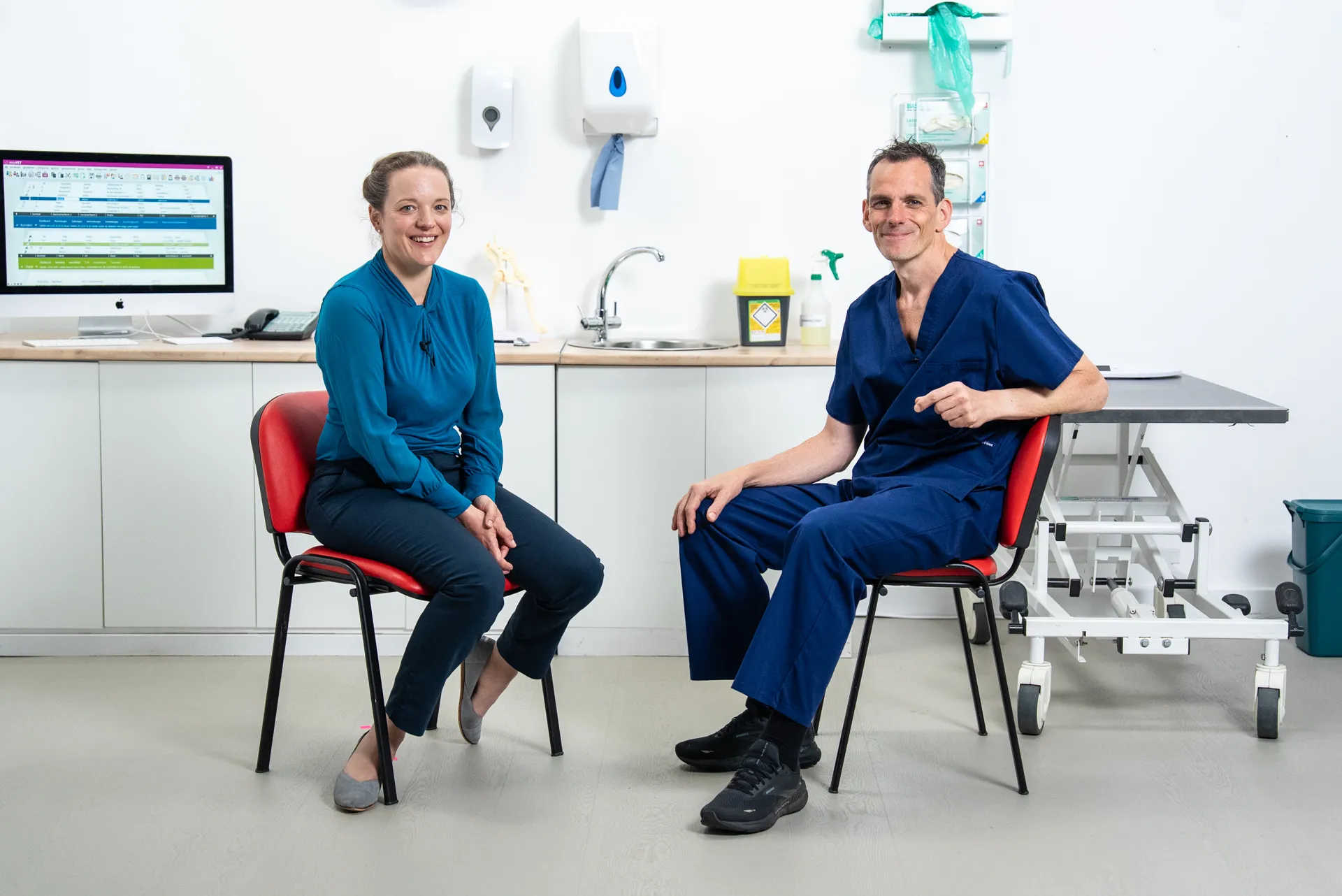Smiling man and woman seated on blue chairs in a modern, well-lit office with plants, bookshelves, and framed pictures.