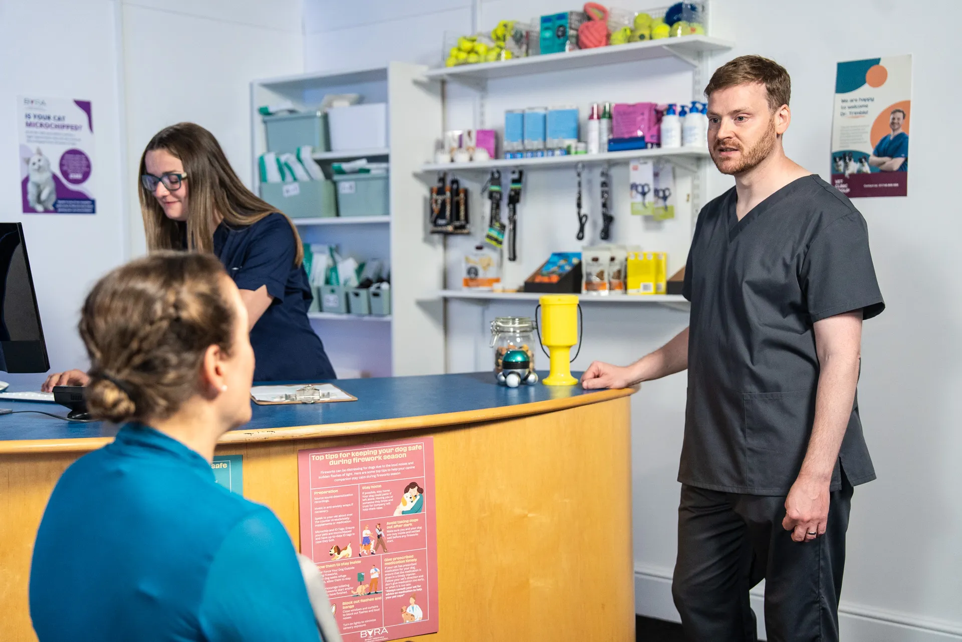 A male veterinarian in black scrubs speaks with a seated female client in a veterinary clinic reception area where a female staff member works.