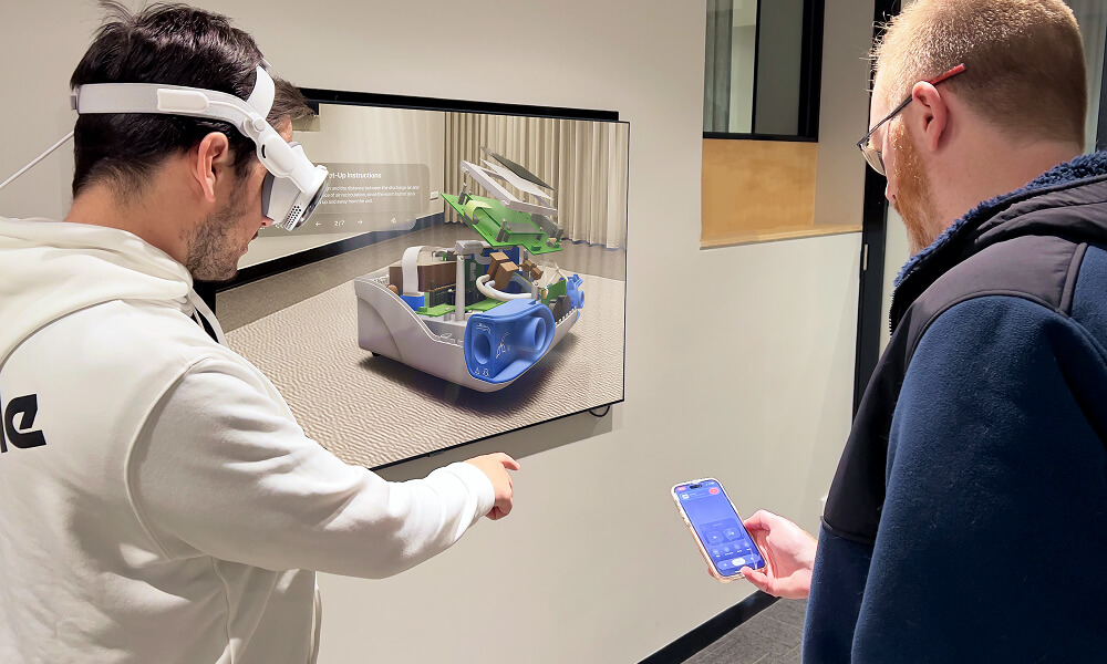 Two men interacting with augmented reality; one wearing a VR headset pointing at a 3D model displayed on a screen, the other holding a smartphone.