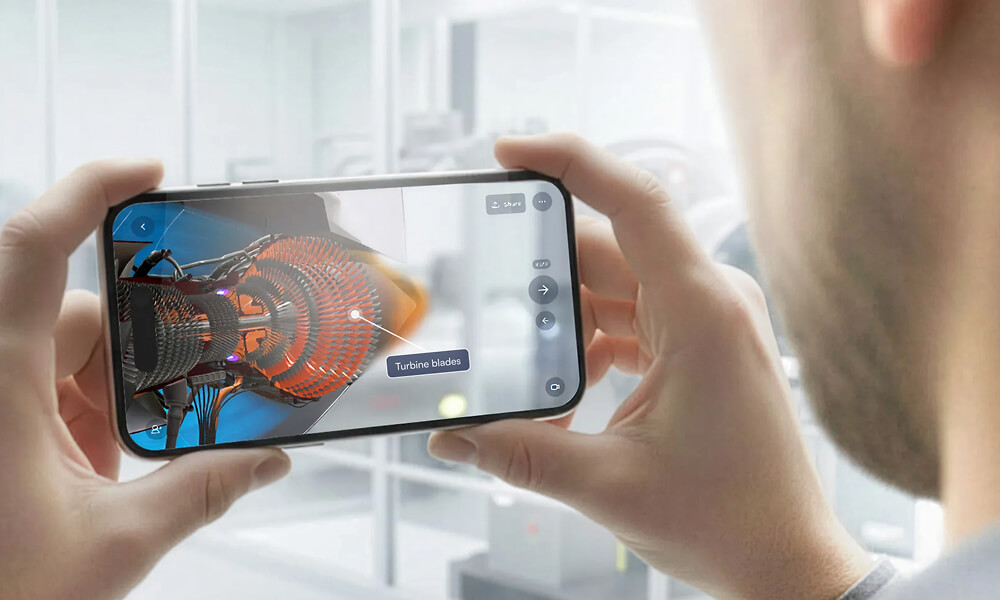 Two men interacting with augmented reality; one wears a VR headset pointing at a 3D mechanical model on a wall screen while the other holds a smartphone.