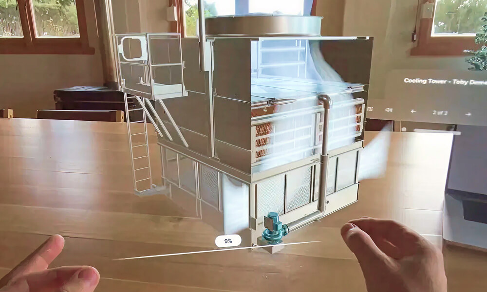 Hands holding a holographic 3D model of an industrial cooling tower above a wooden table.