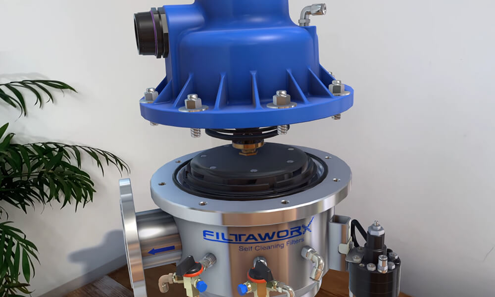 Close-up of a metallic Filtaworx self-cleaning filter with a blue top cover, set against a white wall with a plant in the background.