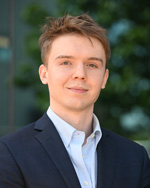 A professional headshot of Andrew Reutsky in a dark navy blue blazer and baby blue shirt, photographed outdoors with a blurred green tree in the background. He is facing the camera with a slight smile.