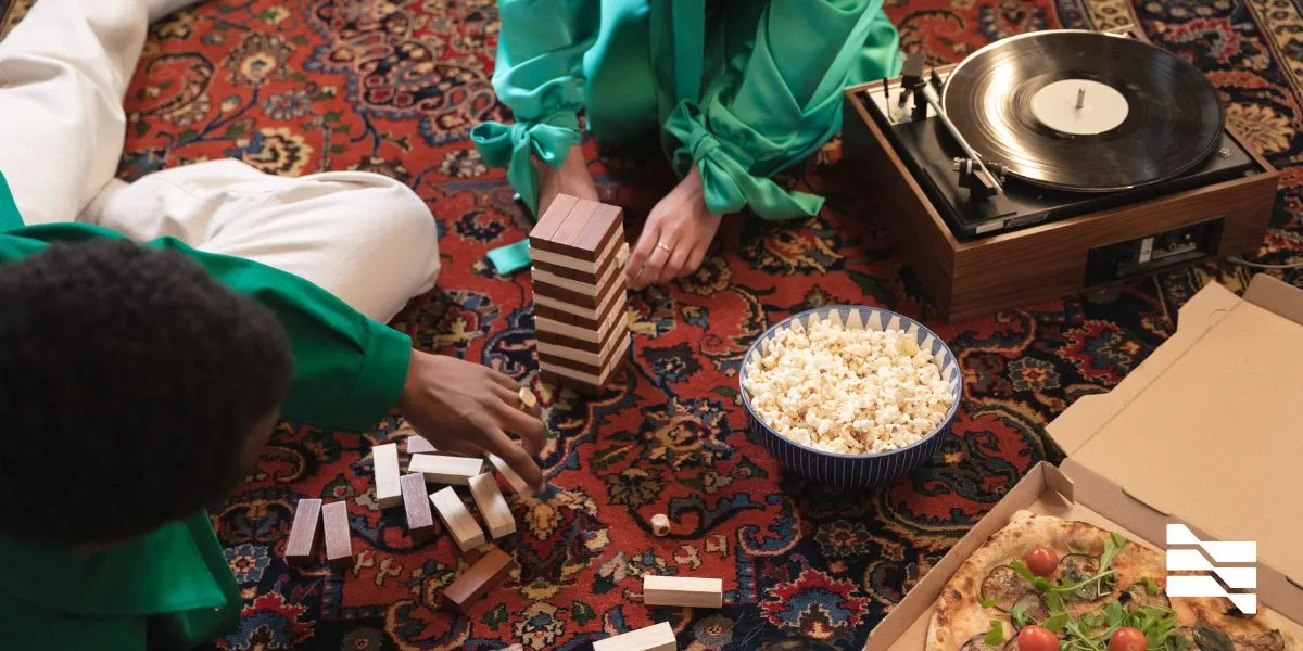 Two people enjoy a homebound retreat with a game of Jenga, vinyl music, popcorn, and pizza, finding joy during COVID-19.