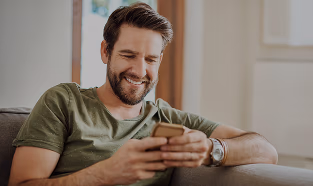 Ein Mann sitzt entspannt auf dem Sofa und schaut lächelnd auf sein Smartphone.