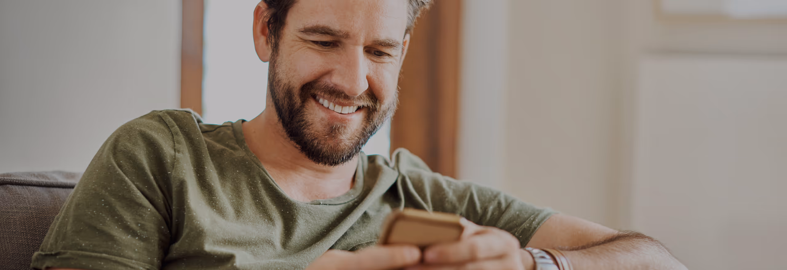 Ein Mann sitzt entspannt auf dem Sofa und schaut lächelnd auf sein Smartphone.