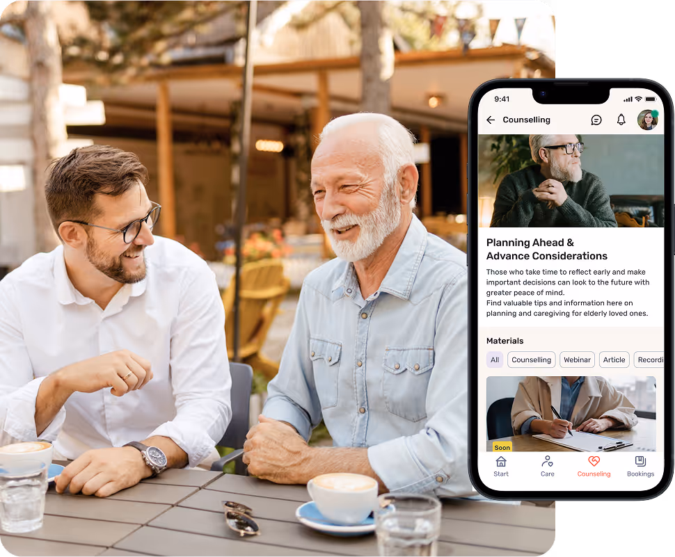 An older man and a younger man are smiling and talking over coffee outdoors. The HeyCare app can also be seen on a smartphone.