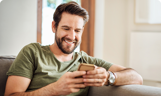 Ein Mann sitzt entspannt auf dem Sofa und schaut lächelnd auf sein Smartphone.