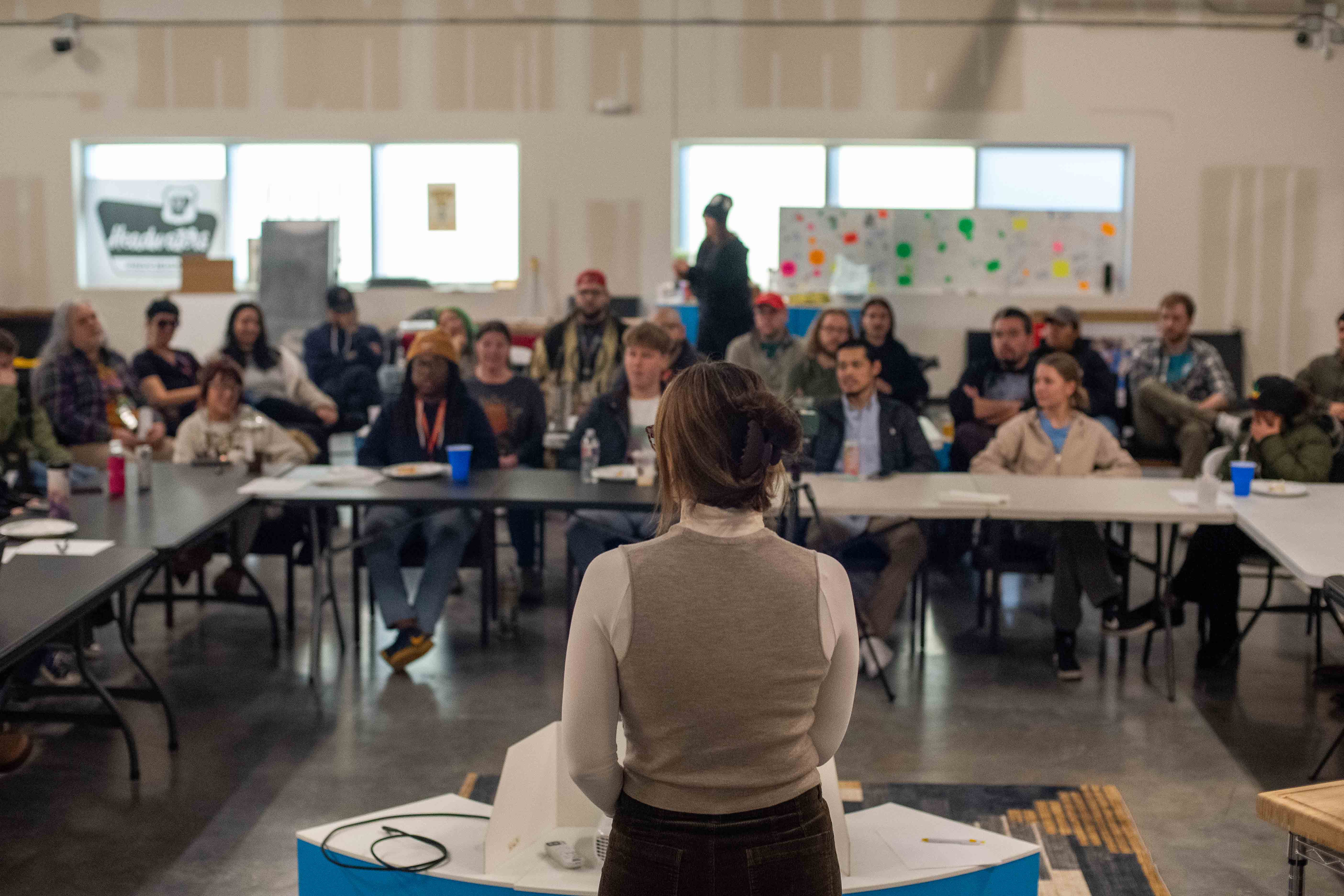Un grupo diverso de personas sentadas en sillas blancas escuchan con atención a un ponente que está fuera de cuadro a la izquierda de la cámara.